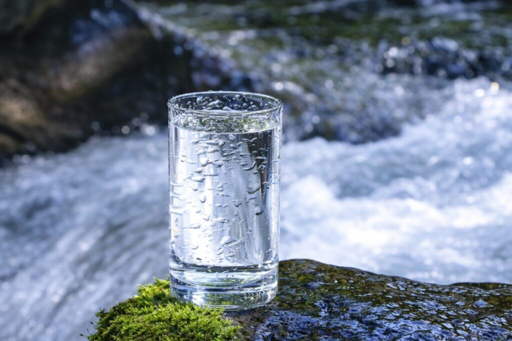 清流のそばの苔むした岩の上に置かれた透明なグラスに入った水と、背景に流れる川の風景