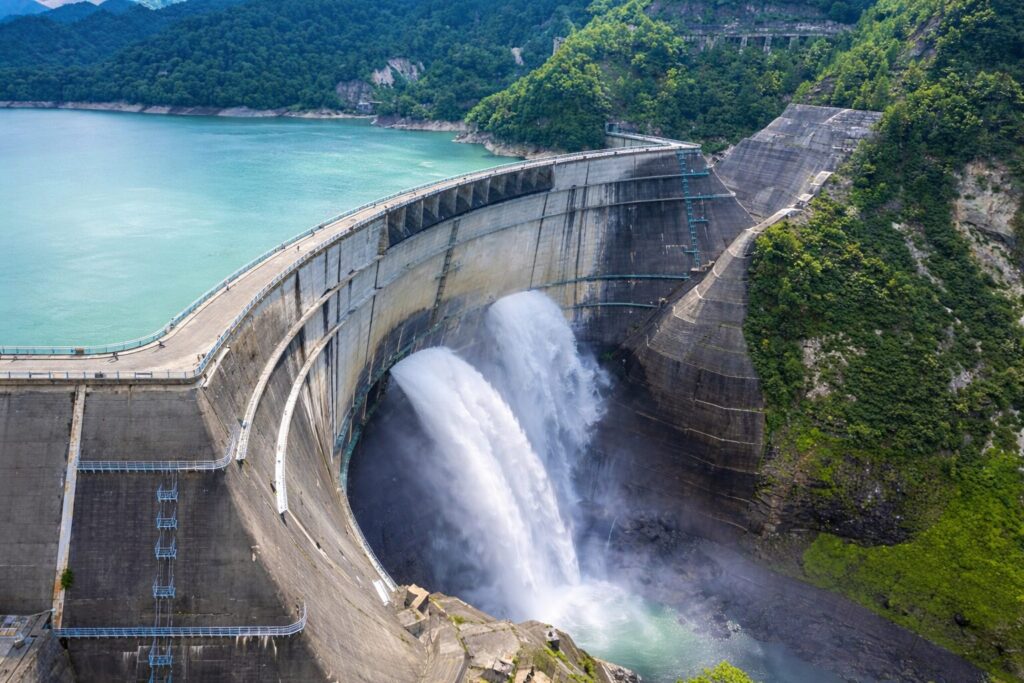 湾曲した巨大なコンクリートダムから勢いよく放水される様子と、エメラルドグリーンの貯水湖、周囲を囲む緑豊かな山々が広がる風景