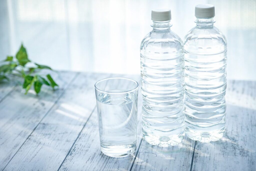 木目のテーブルの上に置かれた透明なペットボトル2本とグラスに注がれた水、自然光が差し込む清潔感のあるシーン