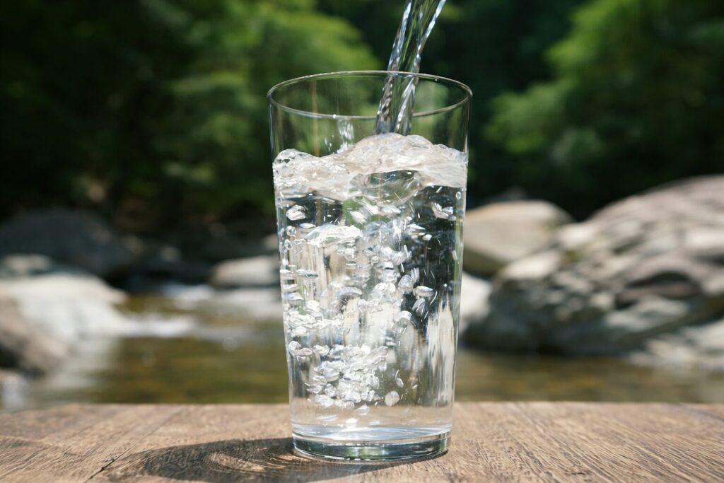 自然の川辺を背景に、透明なグラスへ水が注がれ気泡が立ち上る様子を捉えた清潔感のあるイメージ