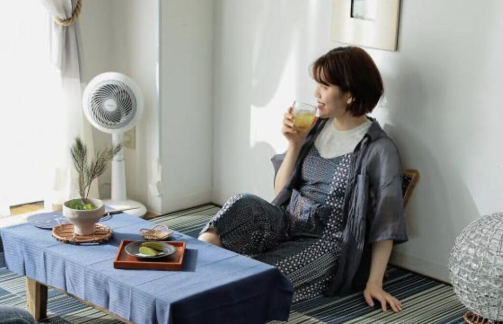 扇風機の風を感じながら室内で飲み物を飲んでくつろぐ女性