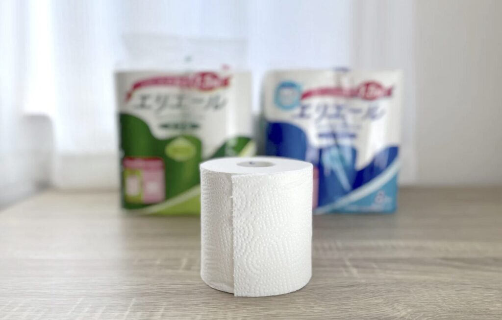 テーブルの上に置かれたトイレットペーパーと後ろに並ぶ家庭用トイレットペーパーのパッケージ