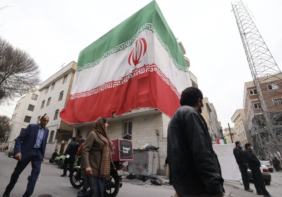 大きなイラン国旗が建物を覆い、街中を歩く人々と中東情勢を象徴する風景