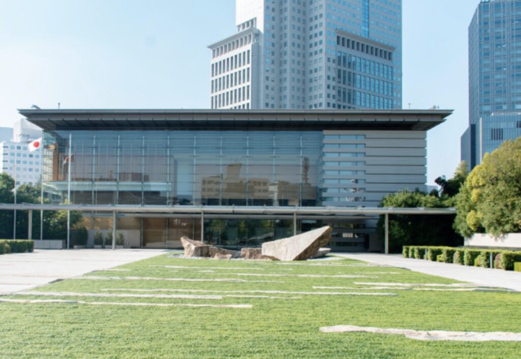 首相官邸の外観全景で、ガラス張りの建物と石造りの広場、日本らしさを象徴する竹や石のデザインが調和した正面玄関の様子