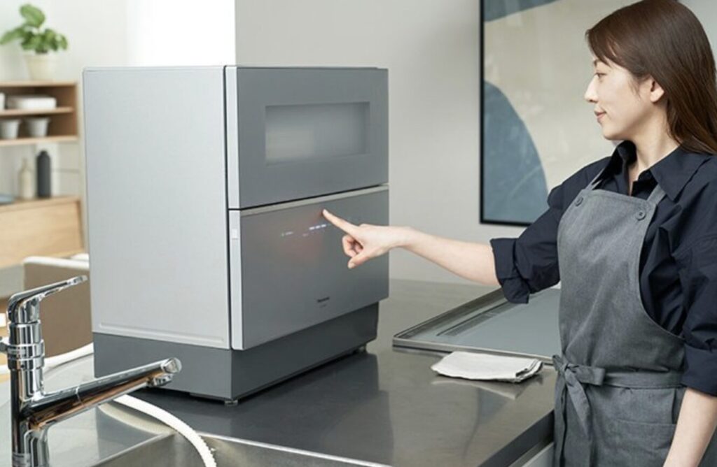 キッチンのカウンター上に置かれた食洗機を操作する女性の様子。シンプルで清潔感のある室内で、タッチパネルに指を伸ばしている。