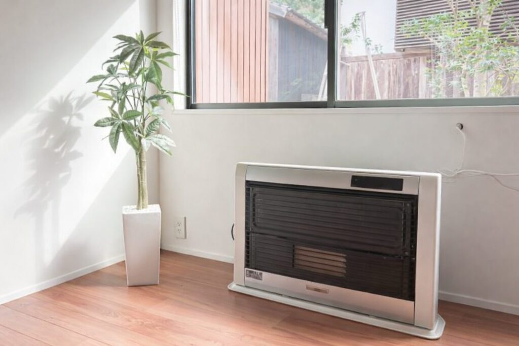 窓から自然光が差し込む室内で、木目の床に置かれた暖房ストーブと観葉植物が並ぶ落ち着いた空間の様子