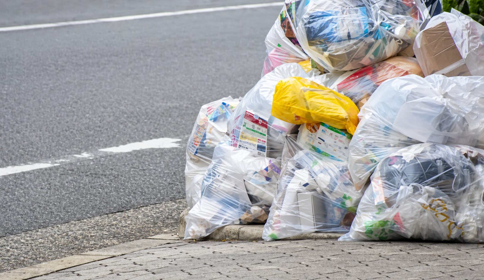 道路脇の歩道に透明なゴミ袋が積み重なり、家庭ごみや包装資材が詰められている様子