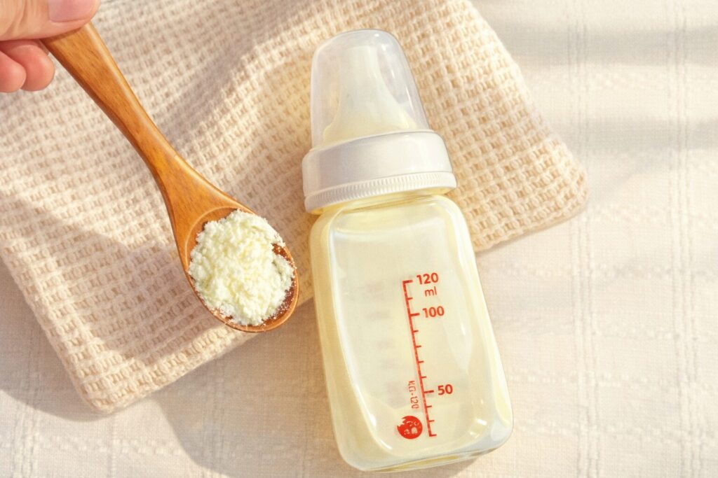 粉ミルクのパウダーを木製スプーンですくい、哺乳瓶と布の上に置かれた様子を描いたやわらかいイラスト