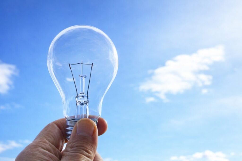 青空と雲を背景に、手で透明な電球を持ち上げている様子のイメージ