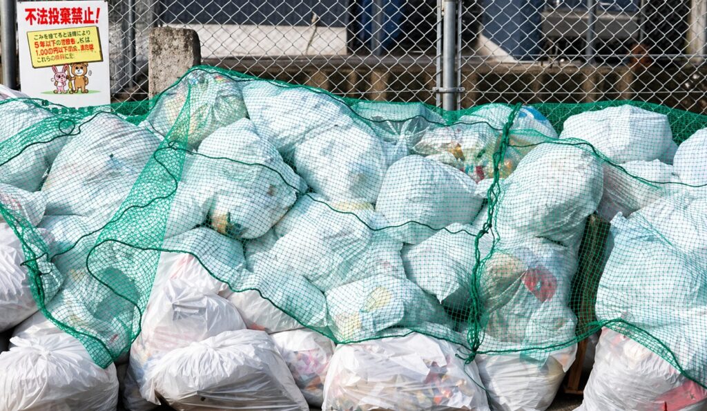 屋外の集積所で白いゴミ袋が山積みになり、緑色のネットで覆われている様子と背後のフェンス