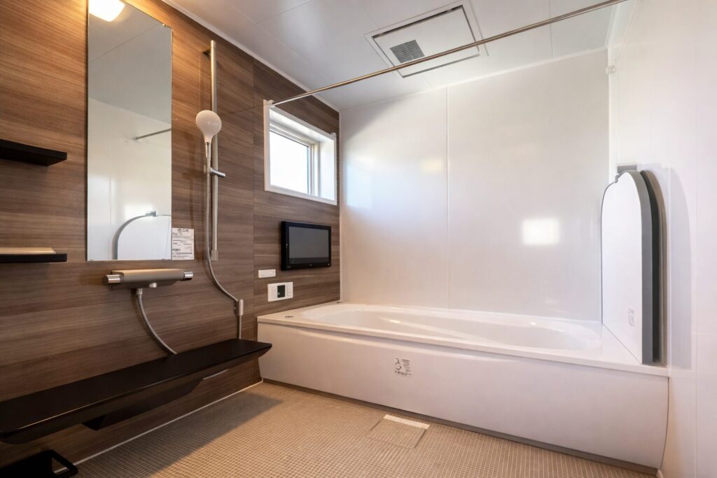 木目調の壁と白い浴槽が組み合わされた清潔感のあるユニットバスで、鏡・シャワー・浴室テレビ・小窓が配置された現代的な浴室空間
