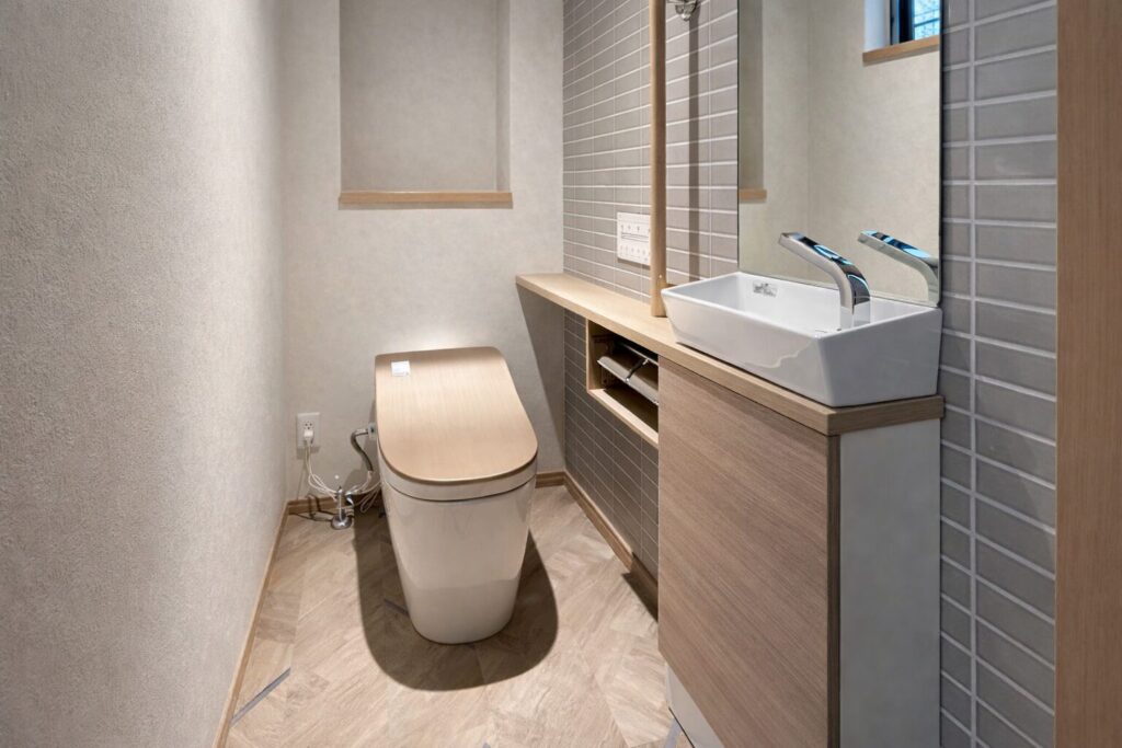 木目調の便座と洗面台が並ぶ、グレーのタイル壁と明るい床材で統一されたシンプルで清潔感のあるトイレ空間