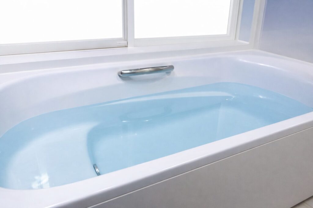 白い浴槽に淡い青色のお湯が張られ、窓からの柔らかい光が差し込むシンプルで清潔感のあるバスルームの様子