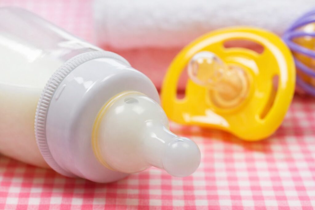 赤ちゃん用の哺乳瓶が横向きに置かれ、奥に黄色のおしゃぶりとラトルがぼかして配置された柔らかい雰囲気の育児用品イメージ