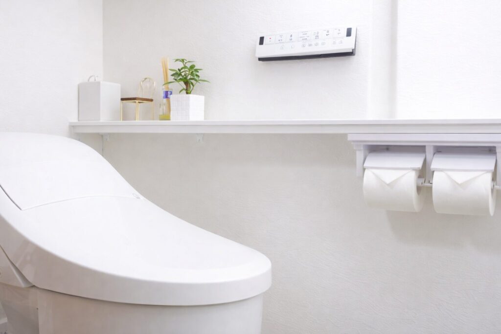 シンプルで清潔感のあるトイレ空間で、左に白い洋式トイレ、右にトイレットペーパーホルダー、壁には操作パネルと棚に植物や小物が配置されたモダンな内装