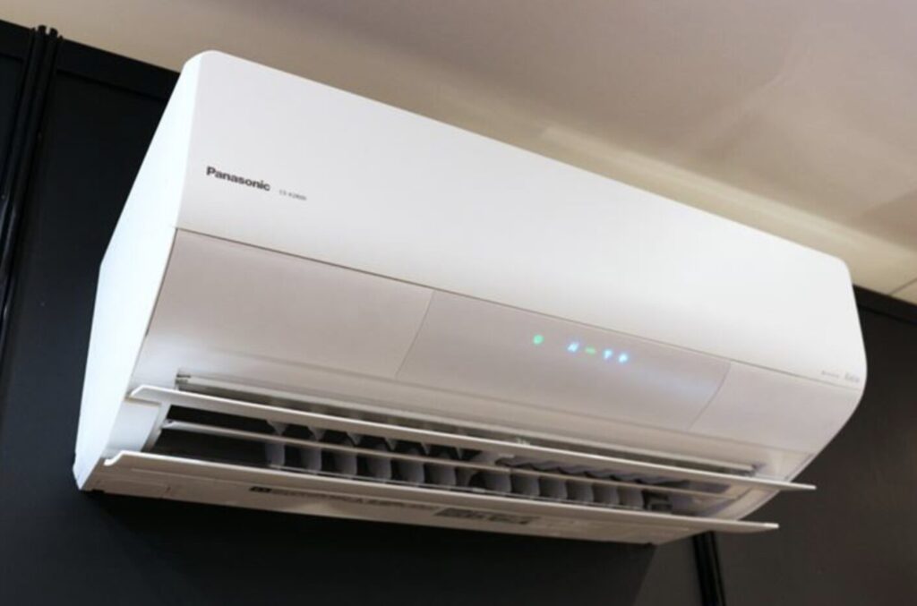 室内の壁上部に設置されたパナソニック製エアコン。前面パネルが開き、内部の送風ルーバーと動作中のランプが点灯している様子。