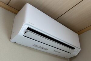 天井付近の壁に設置されたパナソニック製の壁掛けエアコン。吹き出し口のフラップが閉じ気味で、木目の天井とベージュの壁に調和している。