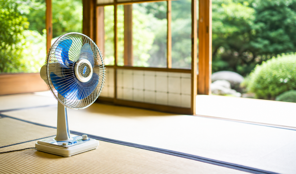 夏の和室の畳に置かれたレトロな青い羽根の扇風機と、窓の外に広がる緑の庭